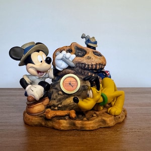 May include: A detailed figurine featuring Mickey Mouse in safari attire, Pluto, and a dinosaur skull. The base is brown with a bone and a circular emblem. Mickey holds a tool, Pluto is on the ground, and a small hammer rests on the skull.