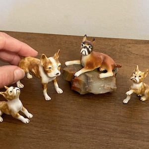 May include: Four porcelain figurines of dogs, two brown and white, one brown and white with black spots, and one brown and white with black ears. The dogs are in various poses, and one is sitting on a rock.