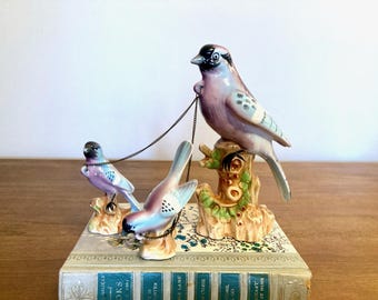 May include: A vintage porcelain bird figurine set with three birds perched on a tree branch. The birds are painted in shades of blue, purple, and white. The birds are connected by a gold chain.