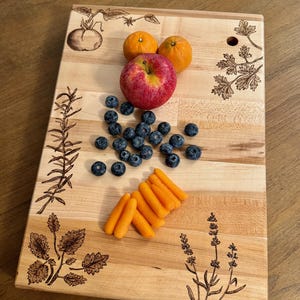 May include: A rectangular wooden cutting board with etched botanical designs and a variety of fresh produce. The board features a red apple, blueberries, orange carrots, and two oranges. The board has a hole in the top right corner.