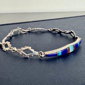 May include: A silver bracelet with a geometric design and a curved section inlaid with colorful stones. The bracelet features a series of interconnected links and a secure clasp. The inlaid section displays blue, turquoise, and other vibrant colors.