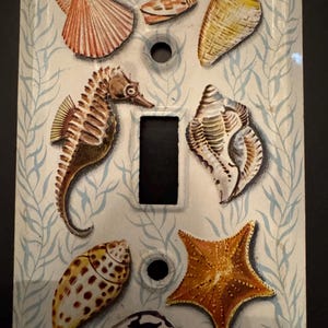 May include: Decorative light switch cover featuring a seahorse, seashells, and a starfish. The design includes a variety of shells in shades of orange, yellow, and white, set against a light blue and white background. The cover has a rectangular opening for the switch.