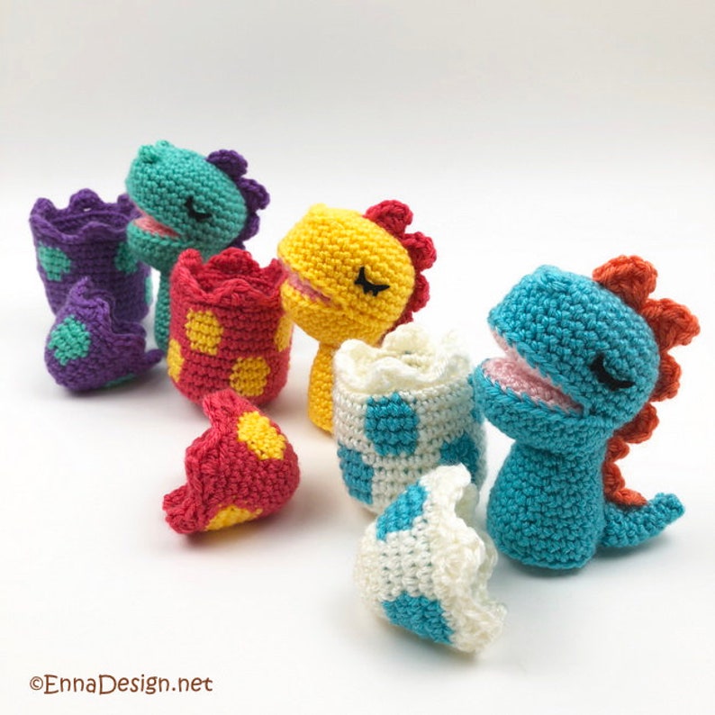 May include: Four crocheted dinosaur toys in different colors, each with a matching egg. The dinosaurs are yellow, blue, green, and purple. The eggs are white with blue, red with yellow, red with yellow, and purple with green polka dots.