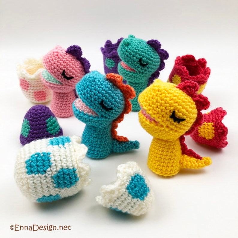 May include: A set of crocheted dinosaur toys in various colors, including pink, blue, yellow, and green. The dinosaurs are in different poses, some are hatching from eggs, and some are standing on their own. The eggs are also crocheted and have a speckled pattern.