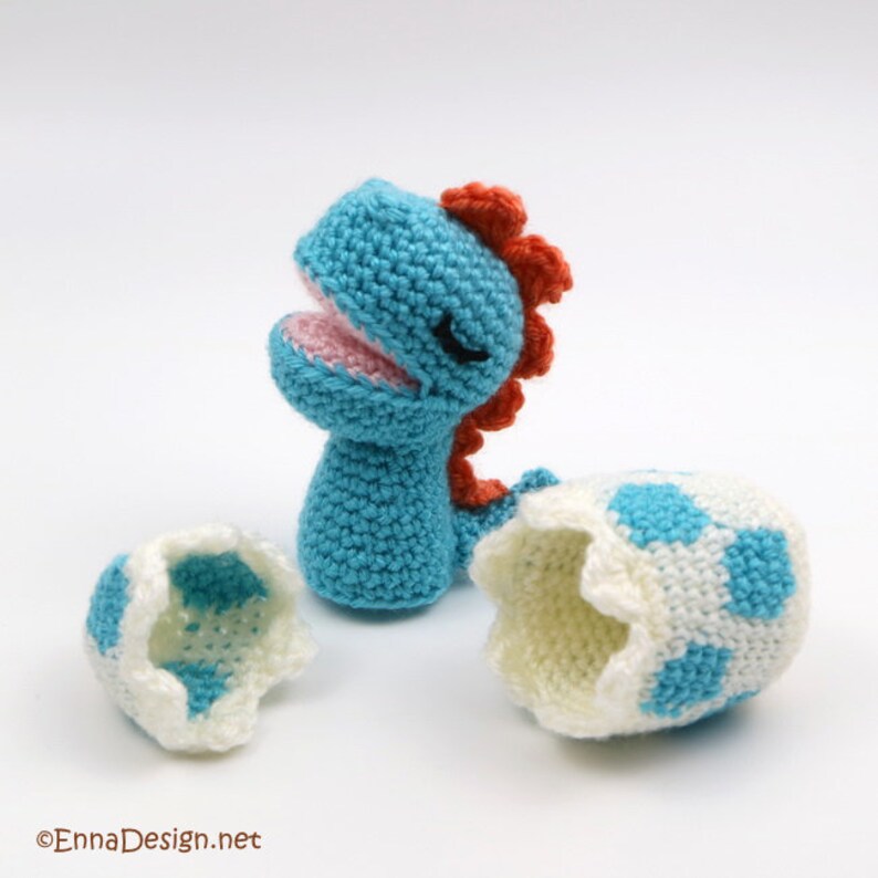 May include: A blue crocheted dinosaur with a red crest emerges from a broken white and blue crocheted egg.