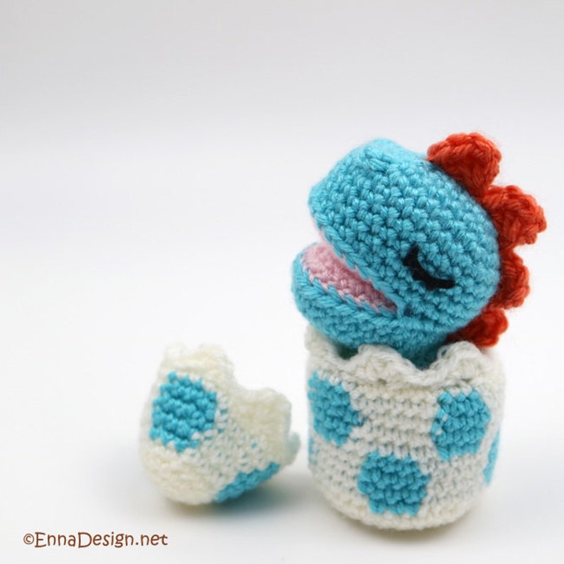 May include: A crocheted blue dinosaur hatching from a white and blue speckled egg. The dinosaur has a pink mouth and closed eyes.