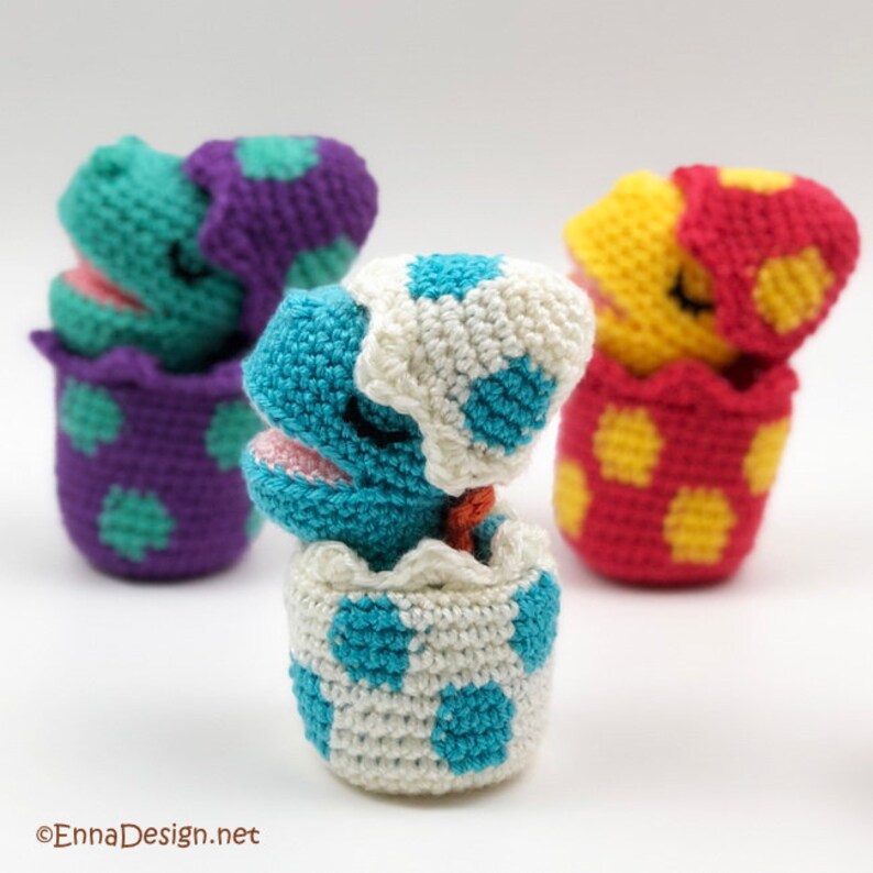 May include: Three crocheted dinosaur toys in eggs. The dinosaurs are blue, purple, and yellow with closed eyes. The eggs are white with blue, purple, and red polka dots.