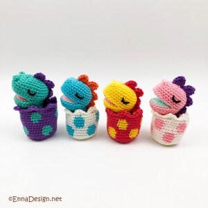 May include: Four crocheted dinosaur toys in different colors, each inside a crocheted egg. The dinosaurs are turquoise, blue, yellow, and pink. The eggs are purple, white, red, and pink.