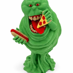 Può includere: Una figura verde e caricaturale con occhi rossi e una bocca larga, che tiene in mano una fetta di pizza e un hot dog. La figura ha un aspetto strutturato, simile a una macchia, con una lingua grande e denti. Su sfondo bianco.