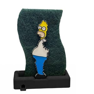 May include: A green, textured sponge with a cartoon image of Homer Simpson. The yellow-skinned character is wearing a white shirt, blue pants, and black shoes. The sponge is set in a black, rectangular holder.