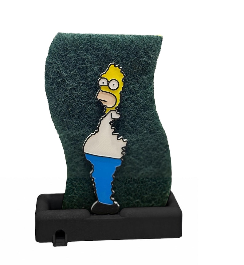 May include: A green, textured sponge with a cartoon image of Homer Simpson. The yellow-skinned character is wearing a white shirt, blue pants, and black shoes. The sponge is set in a black, rectangular holder.