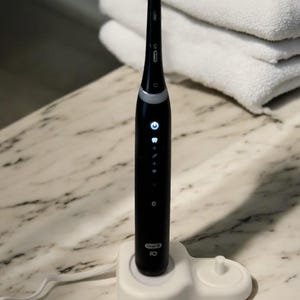 May include: A black electric toothbrush with a white charging base. The toothbrush has a digital display with multiple settings and a blue and white brush head. The base has the text "Oral-B iO" printed on it.
