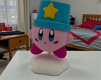 May include: A 3D-printed figure of a pink character with large blue eyes, pink cheeks, and small pink feet. The character wears a blue hat with a yellow star. It stands on a white cloud-shaped base.