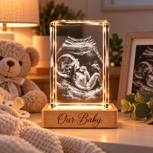 Può includere: Un blocco di vetro rettangolare con un'immagine ecografica di un feto incisa al laser in 3D. Il vetro poggia su una base in legno chiaro con la scritta "Our Baby" incisa sul davanti, illuminata da una fonte di luce calda interna.