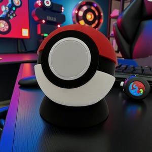 Op de afbeelding: Een rood, zwart en wit bolvormig apparaat met een zilveren midden, dat lijkt op een Poké Ball. Het staat op een zwarte basis op een donker oppervlak. De achtergrond bevat gaming-thema decor en een computermonitor.