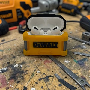 Könnte beinhalten: Gelb-schwarzes Schutzetui für kabellose Ohrhörer mit dem Logo "DeWALT". Das Etui ist geöffnet und zeigt weiße Ohrhörer. Das Etui liegt auf einer Holzoberfläche, im Hintergrund sind Werkzeuge zu sehen.