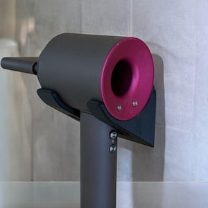 May include: A dark gray hair dryer with a pink nozzle is mounted on a wall with a black bracket. The hair dryer has two buttons on the side.