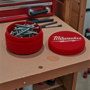 Puede incluir: Un recipiente rojo texturizado lleno de tornillos plateados, junto a su tapa con el logotipo "Milwaukee" en blanco. El recipiente está sobre un banco de trabajo, con una caja de herramientas roja y herramientas en el fondo.