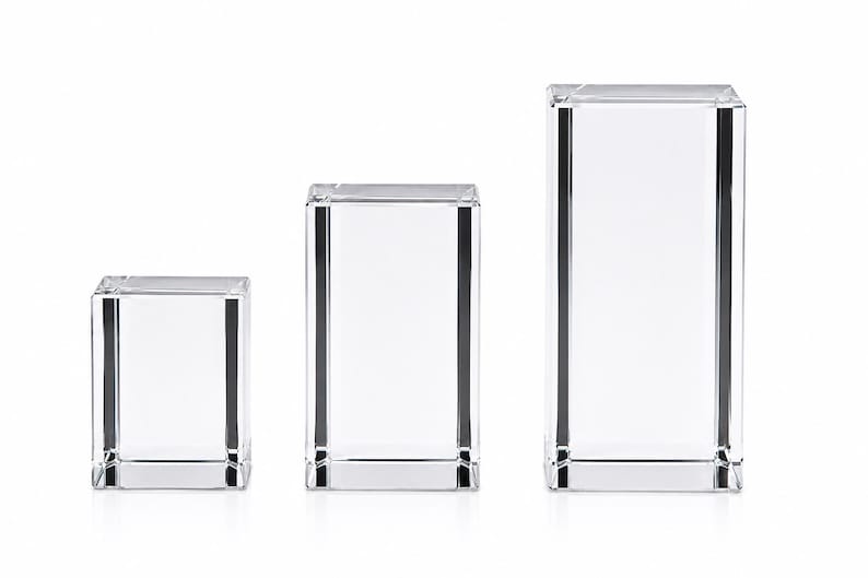 May include: Three clear, rectangular glass display pedestals of varying heights arranged in a row. Each block features a smooth, transparent surface with sharp, defined edges and a solid, heavy appearance, reflecting light through the clear material.