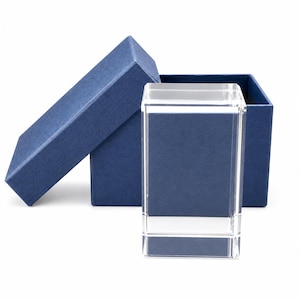 May include: A clear rectangular glass block standing upright in front of an open dark blue textured cardboard gift box. The glass block has sharp, defined edges and a transparent body, while the box features a matching lid resting at an angle.