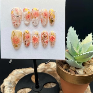 Puede incluir: Un juego de diez uñas artificiales con un diseño floral. Las uñas presentan una base transparente con detalles en rosa, blanco y dorado. Algunas uñas tienen un diseño de flores en 3D. Las uñas se muestran en una tarjeta blanca.