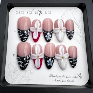 May include: A set of ten artificial nails in a black tray. The nails feature a pink base with various designs, including black and white patterns, white floral accents, and red tips. The text "Thank you for your order. I hope you like it." is also visible.