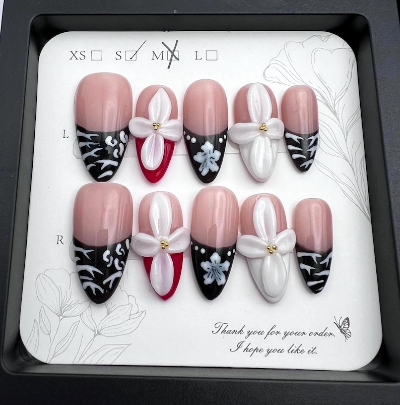 May include: A set of ten artificial nails in a black tray. The nails feature a pink base with various designs, including black and white patterns, white floral accents, and red tips. The text "Thank you for your order. I hope you like it." is also visible.