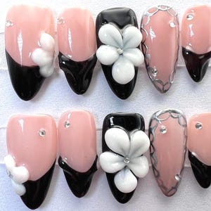 May include: A set of artificial nails with a variety of designs. Some nails are light pink with black tips and white flower accents. Others are black with white flower accents, or light pink with silver trim and rhinestones.