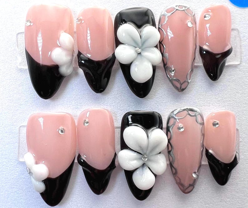 May include: A set of artificial nails with a variety of designs. Some nails are light pink with black tips and white flower accents. Others are black with white flower accents, or light pink with silver trim and rhinestones.