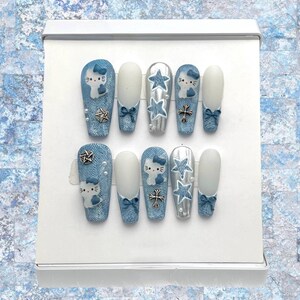 Könnte beinhalten: Ein Set aus zehn künstlichen Nägeln mit einem Jeans-Design. Einige Nägel zeigen weiße Katzenköpfe mit blauen Schleifen, silbernen Sternakzenten und dekorativen Schleifen. Andere Nägel sind einfarbig weiß mit blauen Schleifen.