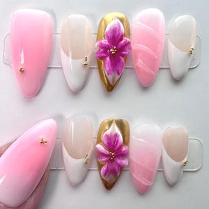 May include: A set of artificial nails in various designs. Some nails are solid pink, others have a white and nude color block design, and some feature a gold base with a 3D pink flower. Small gold studs are also present.