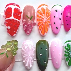 May include: A collection of ten artificial fingernails with various fruit designs. The designs include a red strawberry, pink grapes, an orange slice, a watermelon, and a pink watermelon. Other designs include a green cucumber, a pink nail with white dots, and a green lime.