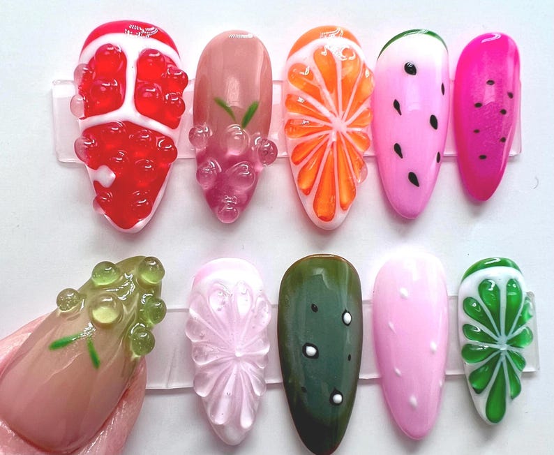 May include: A collection of ten artificial fingernails with various fruit designs. The designs include a red strawberry, pink grapes, an orange slice, a watermelon, and a pink watermelon. Other designs include a green cucumber, a pink nail with white dots, and a green lime.