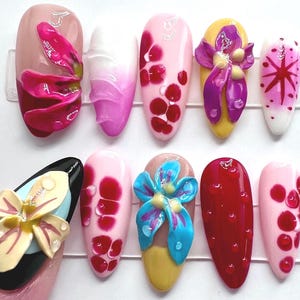May include: A collection of ten artificial nails with various designs. The nails feature a range of colors, including pink, red, yellow, and black. Some nails have 3D floral embellishments, while others have abstract patterns and droplet details.