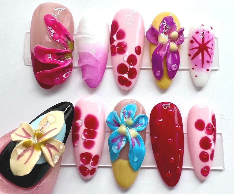 May include: A collection of ten artificial nails with various designs. The nails feature a range of colors, including pink, red, yellow, and black. Some nails have 3D floral embellishments, while others have abstract patterns and droplet details.