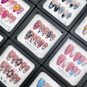 May include: A collection of artificial nails in black display boxes. The nails feature various designs, including floral patterns, animal prints, and abstract art. Some nails have rhinestones and other embellishments. The text "NAIL ART" is visible on one box.
