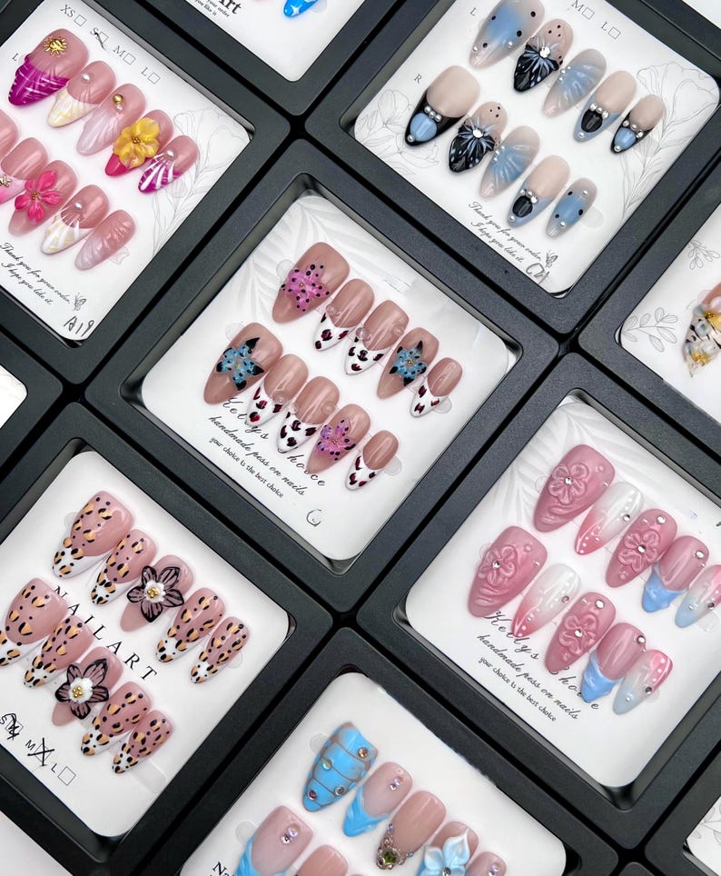 May include: A collection of artificial nails in black display boxes. The nails feature various designs, including floral patterns, animal prints, and abstract art. Some nails have rhinestones and other embellishments. The text "NAIL ART" is visible on one box.