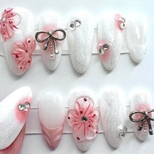 May include: A set of white and pink artificial nails. Some nails feature 3D pink flowers, silver bows, and rhinestones. The nails are oval-shaped and have a glittery finish, with a pink gradient at the base.