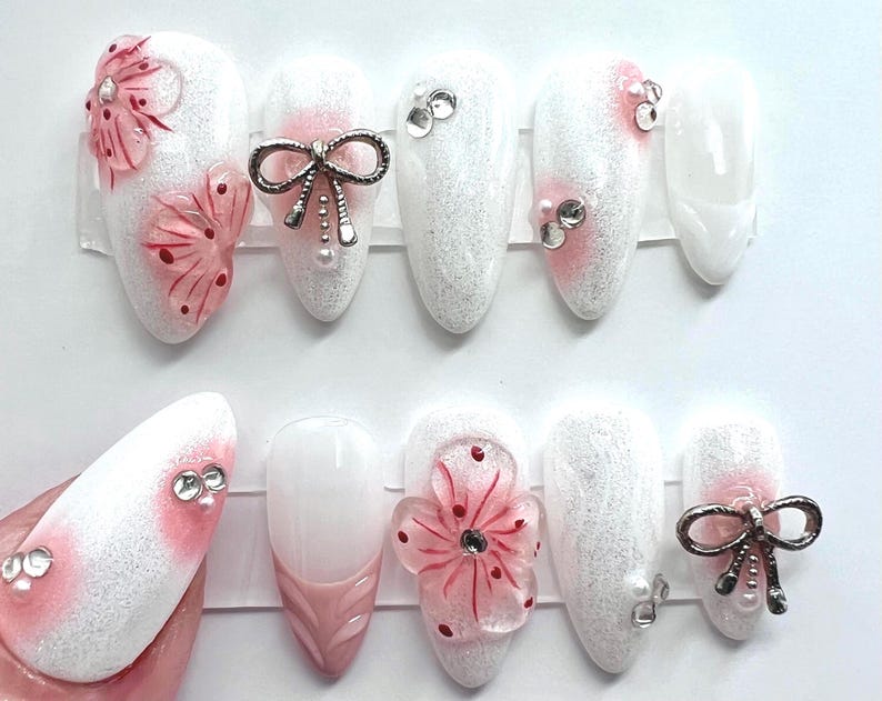 May include: A set of white and pink artificial nails. Some nails feature 3D pink flowers, silver bows, and rhinestones. The nails are oval-shaped and have a glittery finish, with a pink gradient at the base.