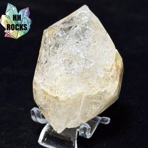 May include: A clear quartz crystal with a pointed top and a rough, textured surface. The crystal is displayed on a clear plastic stand against a black background. The logo "HH ROCKS" is in the upper left corner.