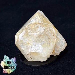 May include: A faceted, light-yellow crystal with a pyramidal shape, resting on a black circular base. The crystal has a translucent quality, with visible internal structures. The logo "HH ROCKS" is in the lower left corner.