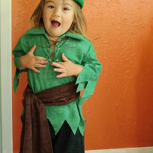 May include: A child's costume featuring a green tunic and hat with a red feather. The outfit includes dark green pants, a brown sash, and a lace-up neckline. The costume is designed for a playful, whimsical look.