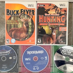May include: Two Wii game cases: "Buck Fever" with a deer and "North American Hunting Extravaganza" with deer, bear, and birds. Below are three game discs: "Spectrobes Origins", "Rock Band", and "Tiger Woods PGA Tour 09 All-Play".