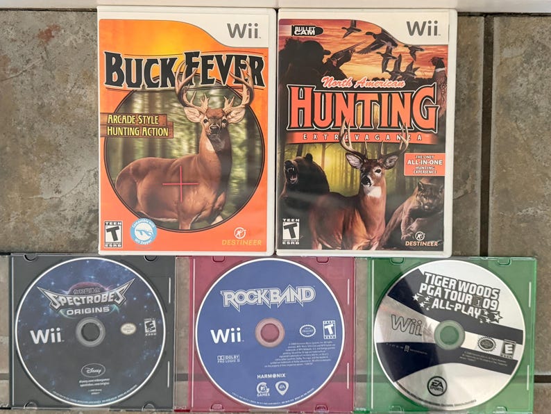May include: Two Wii game cases: "Buck Fever" with a deer and "North American Hunting Extravaganza" with deer, bear, and birds. Below are three game discs: "Spectrobes Origins", "Rock Band", and "Tiger Woods PGA Tour 09 All-Play".