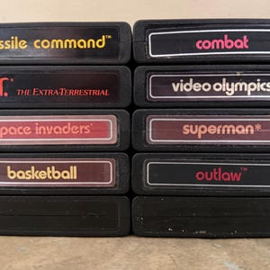 May include: A collection of black Atari game cartridges. Titles include "Missile Command," "Combat," "E.T. The Extra-Terrestrial," "Video Olympics," "Space Invaders," "Superman," "Basketball," and "Outlaw." The text is in various colors.