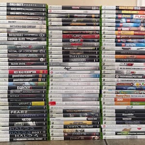 May include: A large collection of Xbox 360 video game cases stacked in three vertical columns. The spines of the cases display various game titles including Call of Duty, Assassin's Creed, Halo, and Borderlands, with consistent green branding.