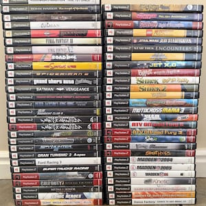 May include: Two vertical stacks of PlayStation 2 video game cases. The spines display various titles including Nobunaga's Ambition, Final Fantasy, Grand Theft Auto, Madden, and Sonic Heroes, featuring diverse color schemes and logos on each case.