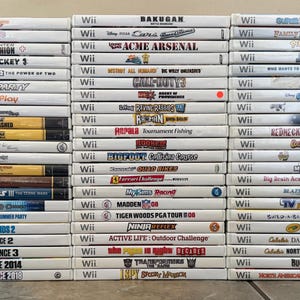 May include: A stack of Wii video game cases, featuring titles like "Just Dance," "Star Wars," and "Madden 08." The cases are white with game titles and logos printed on the spines. The games are stacked on a light-colored surface.