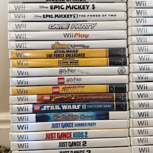 May include: A stack of Wii game cases, featuring titles like "Just Dance," "Epic Mickey," and "Star Wars." The cases are white with game titles printed in various colors. The games are stacked vertically.