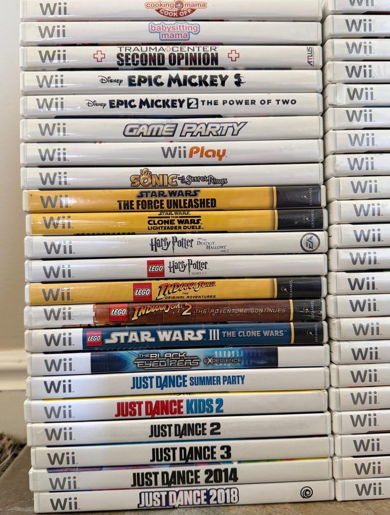 May include: A stack of Wii game cases, featuring titles like "Just Dance," "Epic Mickey," and "Star Wars." The cases are white with game titles printed in various colors. The games are stacked vertically.